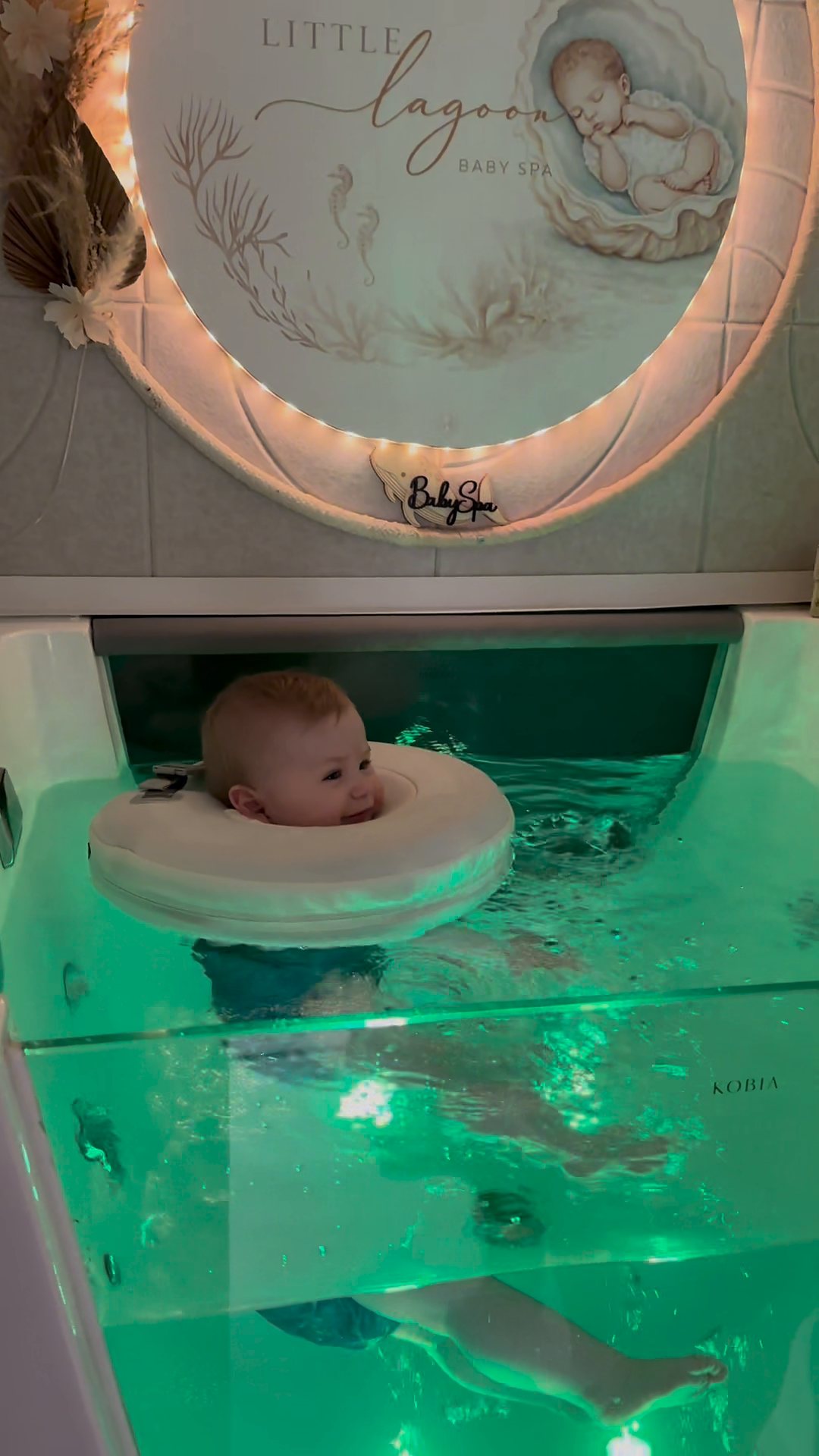 Ein Baby schaut entspannt aus seinem Schwimmring im sanft grün beleuchteten Floating-Becken, darüber das leuchtende Little-Lagoon-Schild