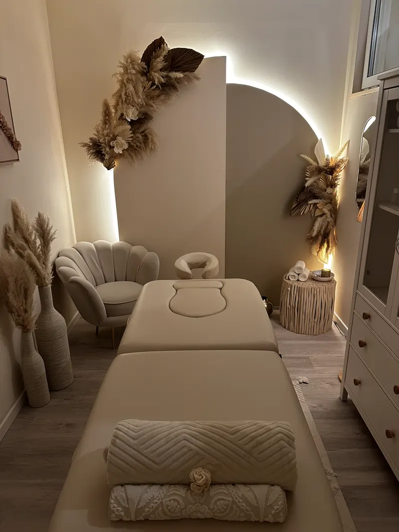 Der warm beleuchtete Massageraum mit Liege, Trockenblumen und Bogenwand